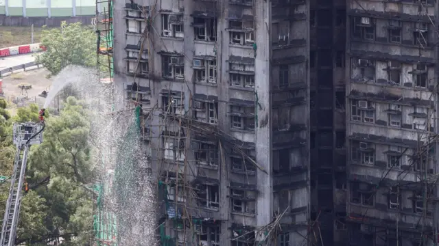 A wide shot of a burnt building with a firefighter on a crane spraying water into it