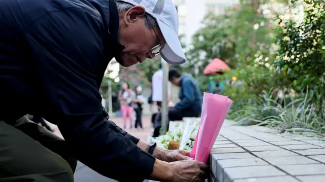 A man laying down a single flower wrapped in pink paper at a makeshift memorial
