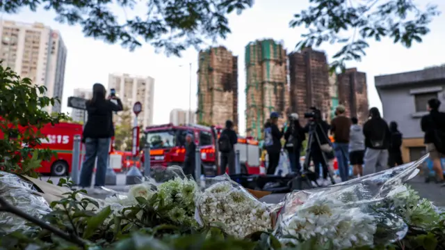 A close up shot of flowers on the ground with burnt buildings in the background