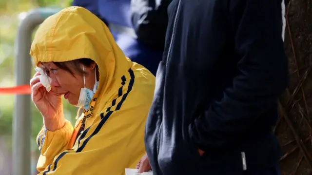 A woman in a yellow hoodie wipes her tears with a piece of tissue paper