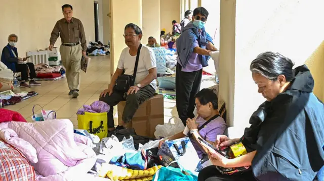 People sitting on boxes and blankets on the ground in a shelter