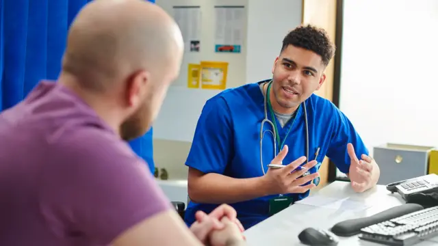 A male clinician wearing blue scrubs speaks to a male patient in a medical clinic