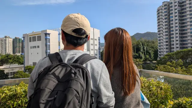 The backs of Mr and Mrs Chan. On the left, Mr Chan wears a grey sweater and a deep-grey backpack; Mrs Chan, with long brown hair, wears a brown top
