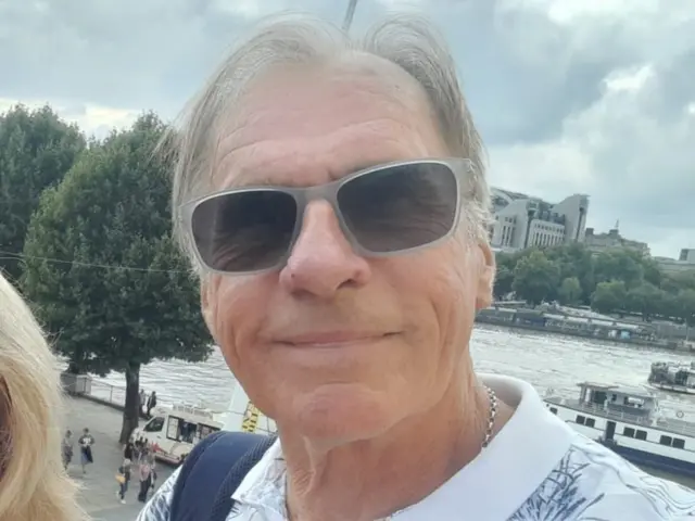 A man wearing grey glasses with trees and buildings behind him