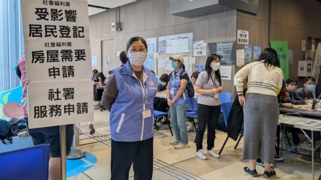A woman with a facemak stands looking into the lense. She is volunteering at a donation centre.