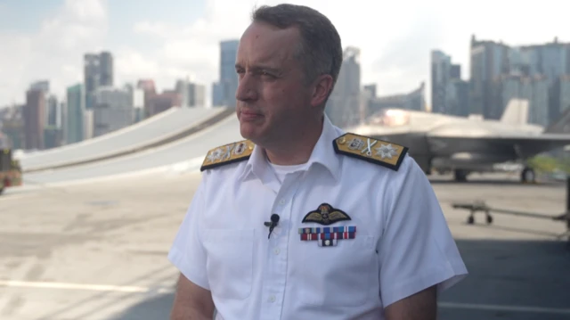 Commodore James Blackmore wears a white short sleeved uniform shirt with black and gold panels on his shoulders. He stands on a grey aircraft platform.