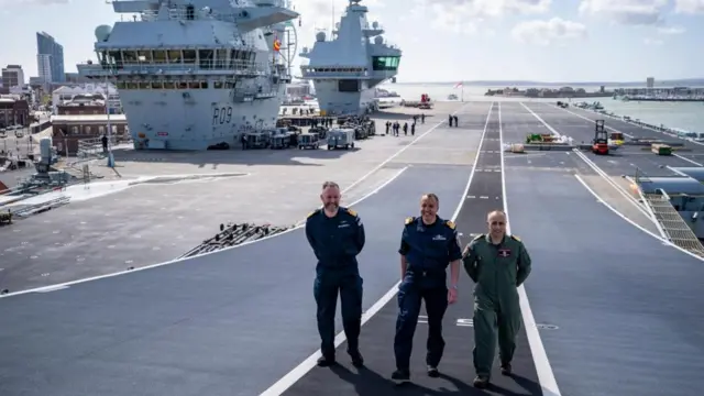 From left to right: Capt Will Blackett, Commodore James Blackmore, and Capt Colin McGannity walk towards the camera wearing dark blue naval uniform and a green flight suit. They are walking alongside the carrier.