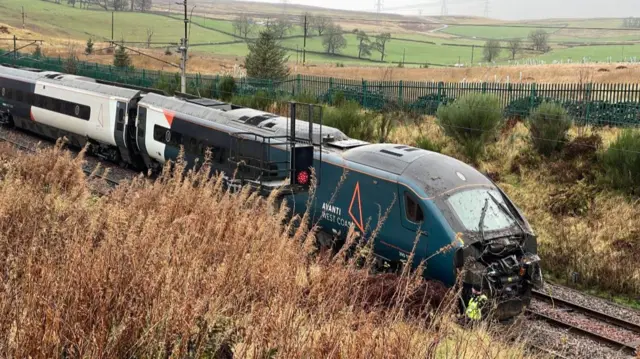 A blue, white and black train damaged at the front is stationary on a railway line in a rural location