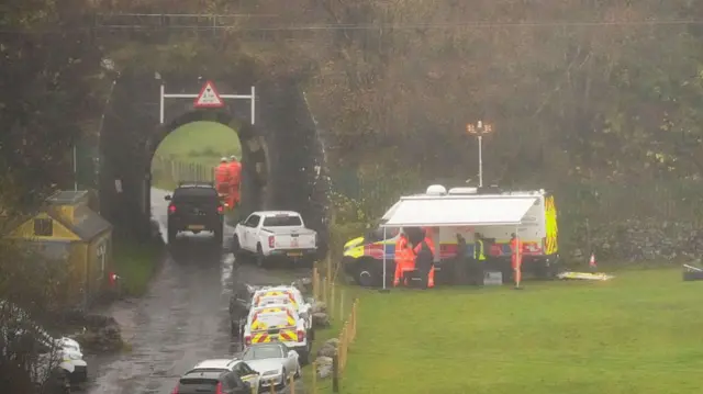 Emergecny service vehicles parked on a road that leads to a small tunnel. There is a tent set up on the right, which is on grass.