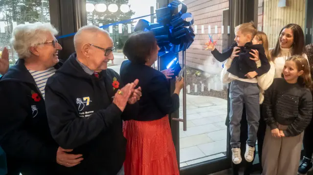 A man and woman with grey hair stand next to a woman with dark hair next to a glass door with a blue ribbon across it. A girl is holding a boy in the air and he is cutting the ribbon with scissors. A girl and a woman with long dark hair stand next to them.