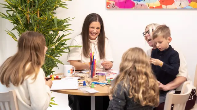 A woman with long dark hair sits at a table next to a woman with blonde hair, a boy and two girls. They are laughing. On the table there is a pot of colourful pens, paper, toys and a mug. Behind them is a colourful image of umbrellas and a plant.