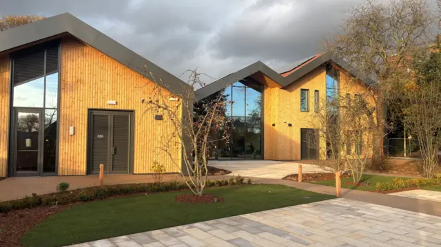 A modern wood clad building with large full height glass windows. A paved area outside the building with grass beds with trees planted in them.