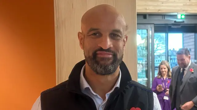 Former Leeds Rhinos player Jamie Jones Buchanan, posing for a photo. He is pictured wearing a dark gillet, with a poppy below his left lapel. An orange wall is behind his right shoulder.