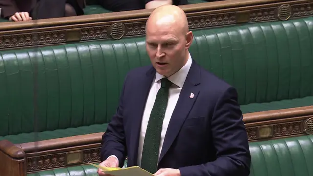 A man in a blue suit and green tie stands in the House of Commons chamber