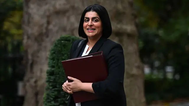 Shabana Mahmood in a black blazer and white shirt, she is holding a red leather folder.