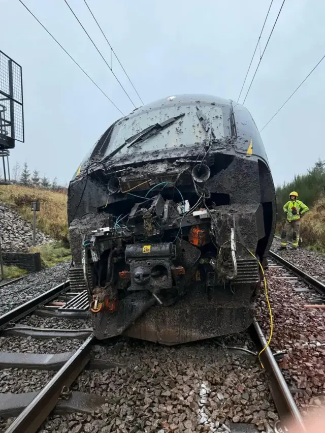 Close up of the front of the train, seen all damaged as it sits on the track. To the right is a person in green hi-vis and a yellow hard hat