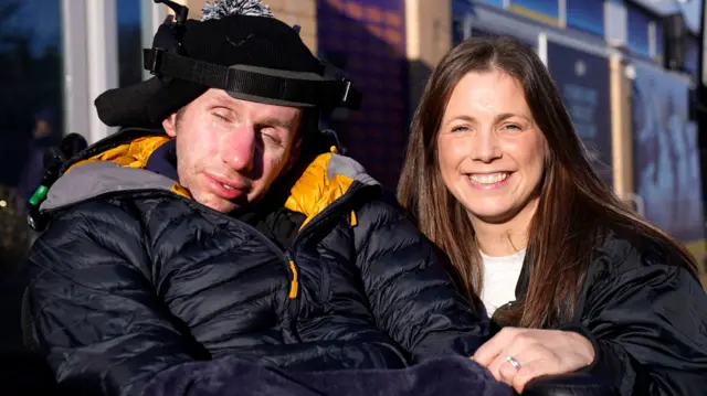 A man in a wheelchair wearing a black and orange puffer jacket with a strap around his forehead next to a woman with long darl hair wearing a black jacket who is smiling at the camera.