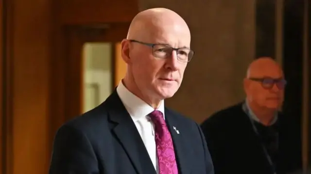 John Swinney, a man wearing a dark-coloured suit, white shirt and purple tie.