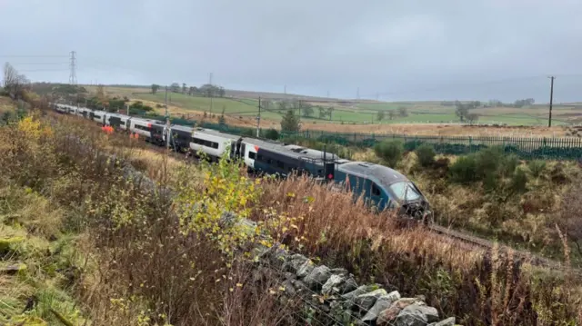 The damaged train on tracks in a rural location with stone wall and grass in the foreground