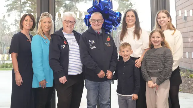 A group of eight people, some of them children, smile and pose for a photo in front of some large glass doors and a blue ribbon.