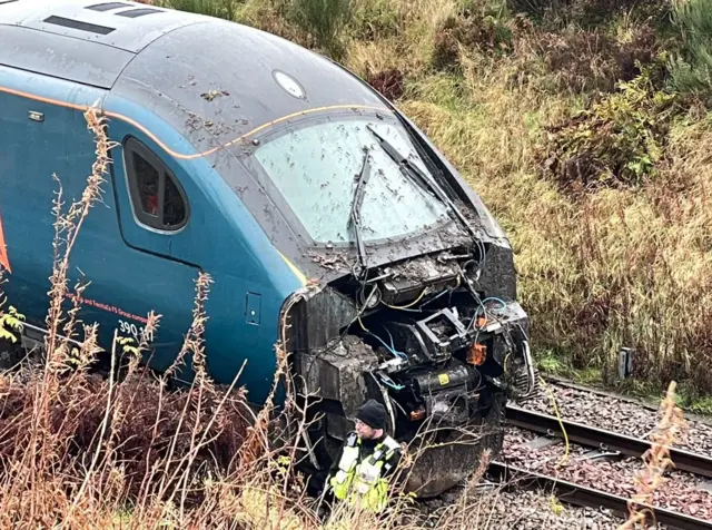 A close-up of the damaged front of a train with a man in hi-vis next to it, on tracks in a rural location