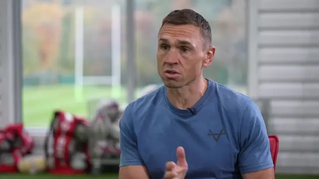A man with short brown hair is wearing a blue t-shirt and gesturing towards the left of the frame. In the background are rugby bags and balls and a window out on to a rugby pitch.