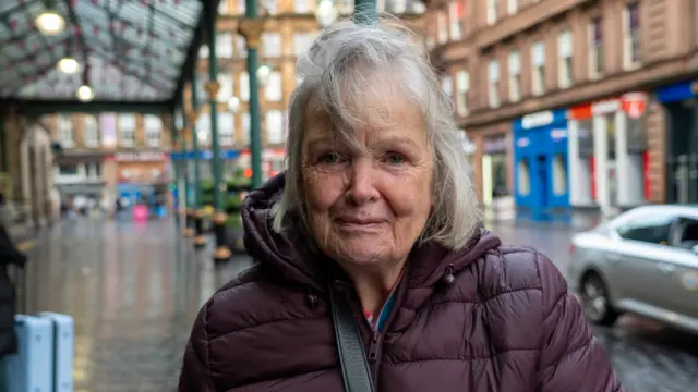 Grandmother Christine Kane stands on the wet street outside Glasgow Central station in a black raincoat