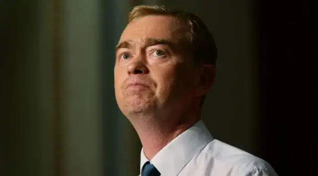 Tim Farron MP, a man in a white shirt and blue tie against a brown backdrop,