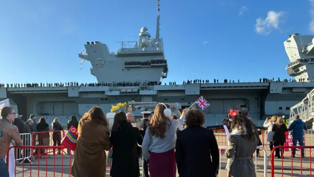 Women are waving Union Jack flags, facing away from the camera and looking at HMS Prince of Wales which is docked