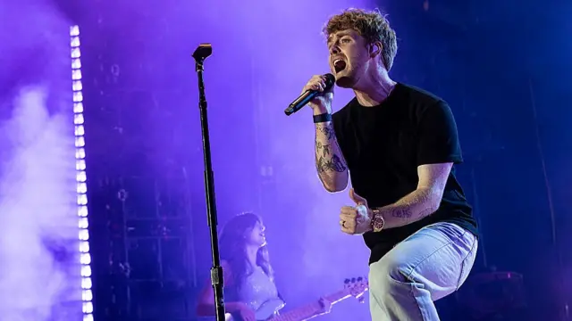 Alex Warren on stage with a mic in his hand, a woman behind him plays bass.