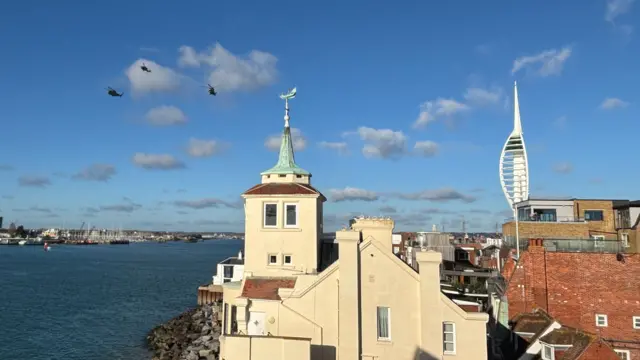 Three helicopters are flying through the sky, it's a bright blue sunny day and the Spinnaker Tower can be seen to the right