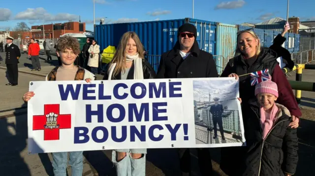 Mabel, Evie and George are all grinning at the camera, standing with their mum, Sammy and another gentleman. They are holding up a sign which says 'Welcome Home Bouncy!'