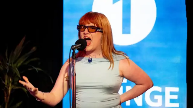 CMAT performing in front of a blue and white digital screen reading Radio 1 live lounge