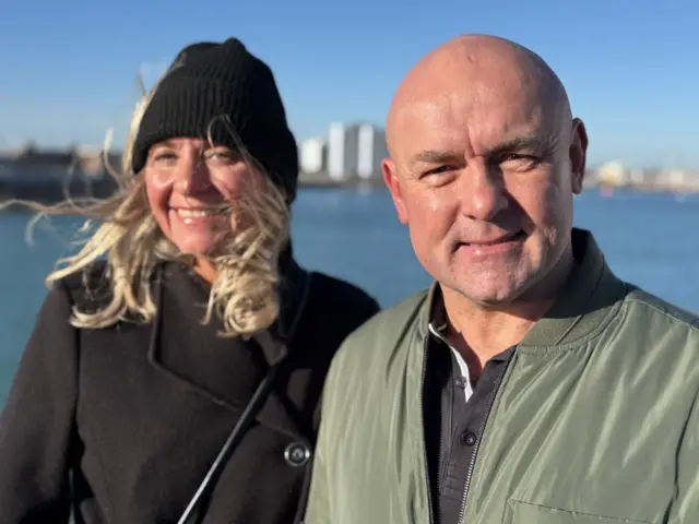 Joanne Chambers is smiling at the camera, she has shoulder length blonde hair which is swept up with the wind under a black knit beanie hat and wears a black smart coat. Gavin Stapleton is bald and smiles at the camera, he wears a green bomber jacket with a black polo top underneath.