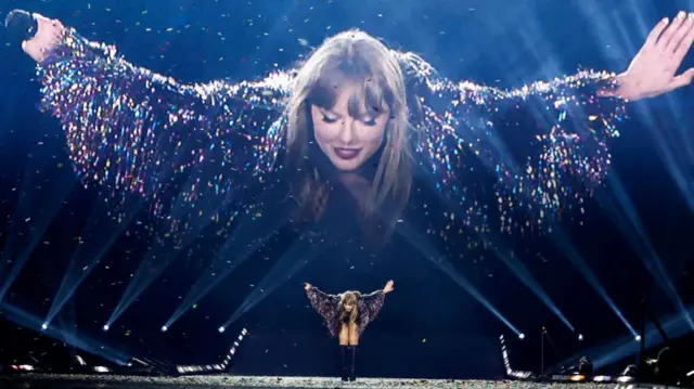 Taylor Swift bowing to the audience, a huge screen behind her shows the same image.