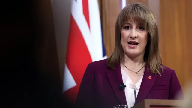 Rachel Reeves stands and gives her speech. We see her and the shadows of reporters in the foreground. She's wearing a burgundy suit with a red poppy.