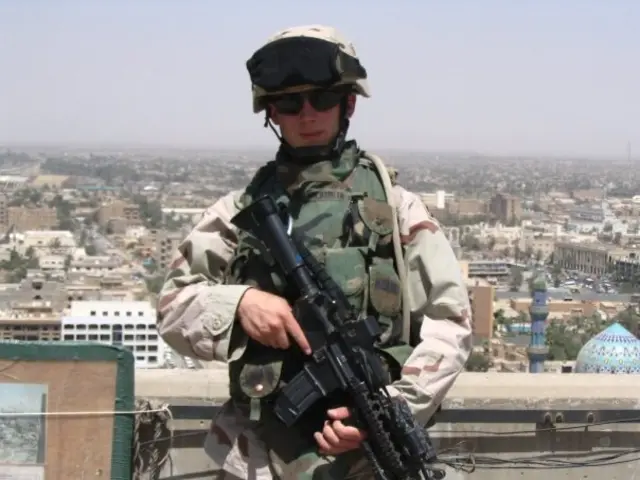 Kristofer Goldsmith in Iraq in 2005. He is wearing US Army uniform and holding a large black gun. Behind him is the backdrop of an Iraqi town.