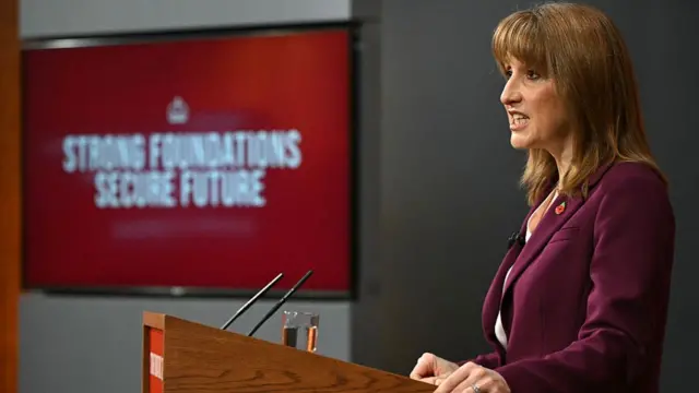Rachel Reeves seen from the side, stood at the pedestal for her speech. She is wearing a burgundy suit with a red poppy badge.