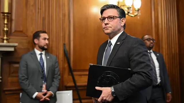 Mike Johnson stands in a wood-panelled room. There are two men stood behind him. He's wearing a black suit and looking seriously outward.