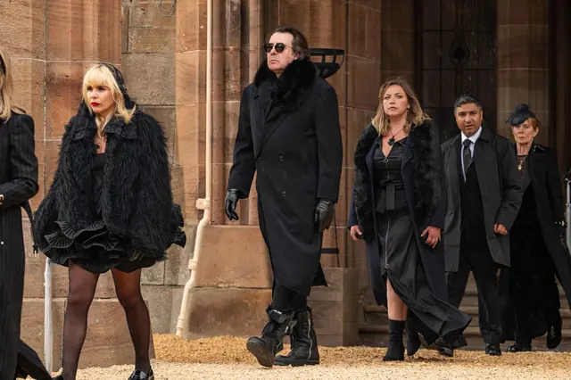 Paloma, Jonathan, Charlotte, Nick and Celia walk outside the castle in black