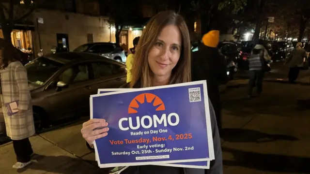 A woman standing on a street at night holds up a blue and white sign which encourages people to vote for Cuomo