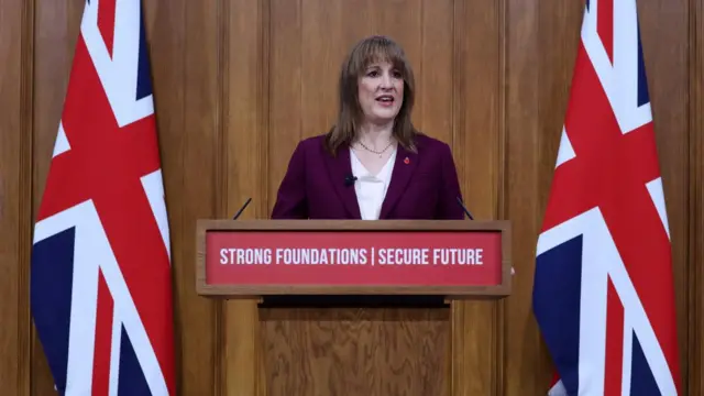 Rachel Reeves stands at a pedestal calmly giving her speech. There are two UK flags on either side of her in the background. Her pedestal says 'Strong Foundations, Secure Future.'