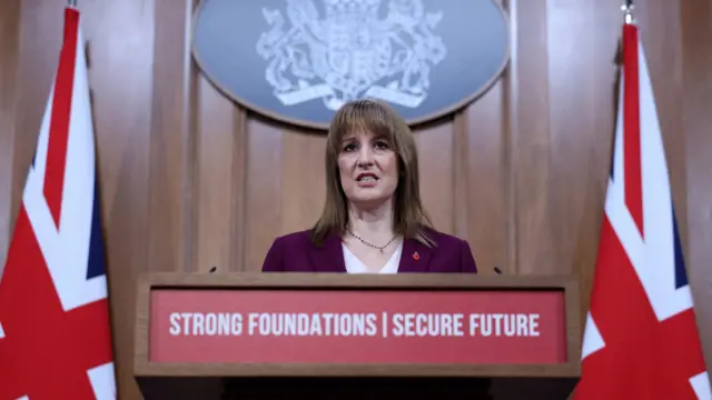 Rachel Reeves stands at a pedestal calmly completing her speech. She has two UK flags behind her and the banner on the pedestal says 'Strong Foundations | Secure Future'. She's wearing a burgundy suit with a poppy badge.