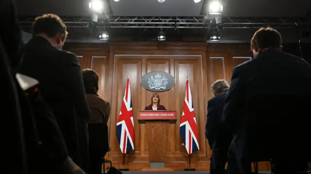 Rachel Reeves stands in front of members of the press, giving her speech. She's stood against a wooden background with two large UK flags.
