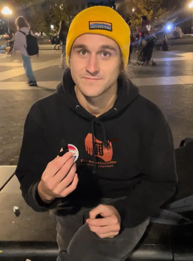 A young man sits on a park bench wearing a black hoodie and a yellow knit hat