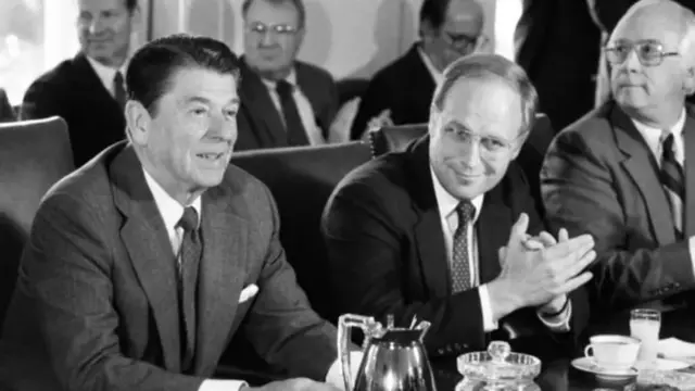 Younger Cheney pictured in black-and-white photograph with former US president Ronald Reagan. Both are sitting at table with coffee pitcher in front of them.