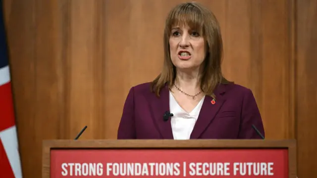 Rachel Reeves speaks from her podium, which says 'Strong Foundations | Secure Future' on it. There's a UK flag in the background. She's wearing a burgundy suit with a red poppy on.