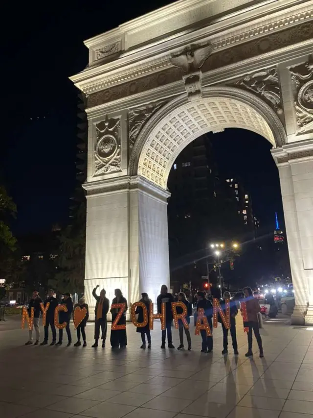 People stand with signs that spell out to read NYC Zohran!, they stand in front of a large archway
