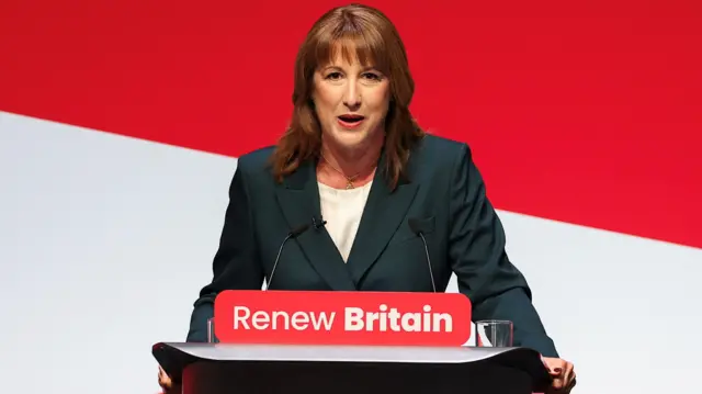 Rachel Reeves delivers a speech. She stands at a lectern on which the words 'Renew Britain' are printed. She wears a grey blazer and white top, and stands in front of a red and white background