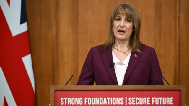 UK Chancellor Rachel Reeves wearing a burgundy suit and remembrance poppy standing at a lectern with the slogan "Strong Foundations: Secure Future"